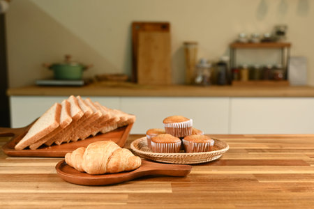 A plate of croissants and muffins sits on a wooden table. The table is surrounded by various kitchen...