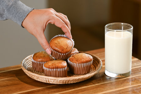 A person is reaching for a muffin on a plate next to a glass of milk. The muffins are arranged in a row on the plate, and the person's hand is almost touching the top of the muffinsの写真素材