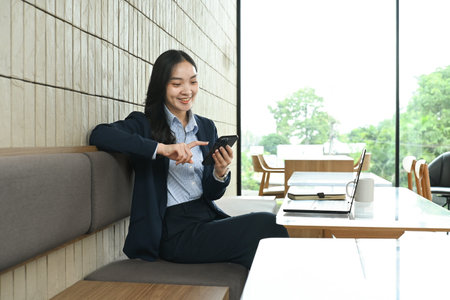 A woman is sitting at a table with a laptop and a cell phone. She is focused on her phone, possibly texting or browsing the internet..の写真素材