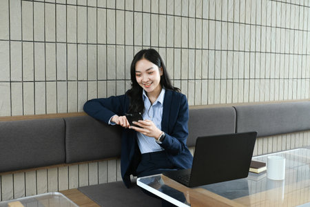 A woman is sitting at a table with a laptop and a cell phone. She is looking at the cell phone.の写真素材