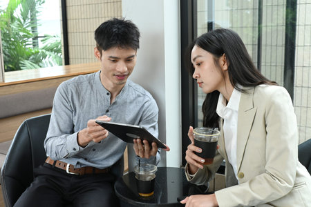 A man and a woman are sitting at a table, looking at a tablet. The man is holding a tablet in his...