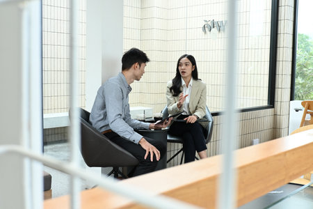 Two people are sitting at a table, one of them holding a tablet. They are talking to each other, possibly discussing work or a project. The atmosphere seems to be professional and focused.の写真素材