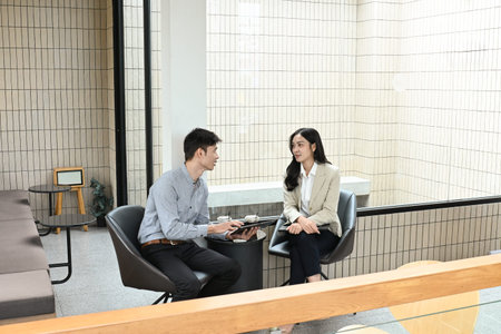 Two people are sitting at a table, one of them holding a tablet. They are talking to each other,...