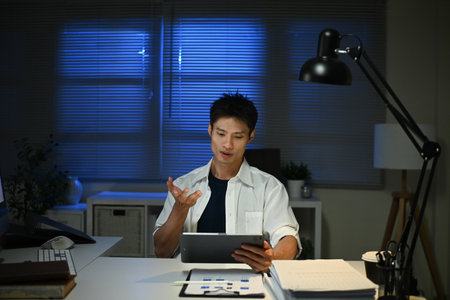 Asian businessman focus online video call meeting assignment on paperwork with colleague in tablet computer in office overtime at night.の写真素材