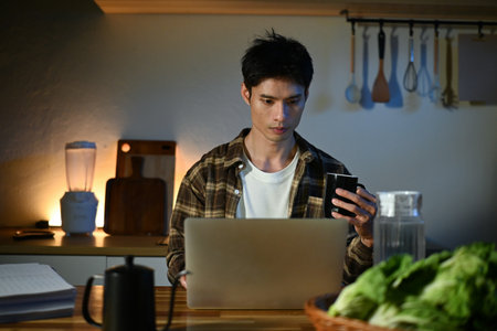 Young man sits in the kitchen at the bar counter works in laptop and drinks a coffee. Home atmosphere, work in the evening after a hard day. Home office.の写真素材