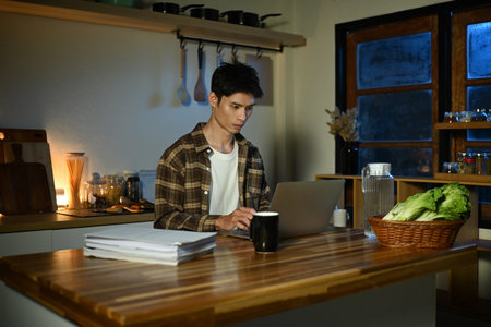 Focused man working late at night on laptop in kitchen with coffee.の写真素材