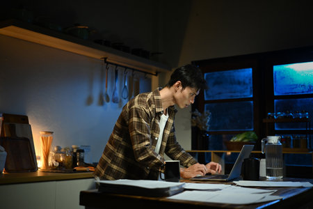 Focused man working late at night on laptop in kitchen with coffee.の写真素材