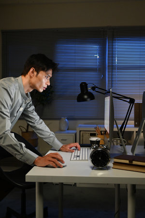 A man is stand at a desk at night with a computer and a keyboard. He is looking at the computer screen.の写真素材