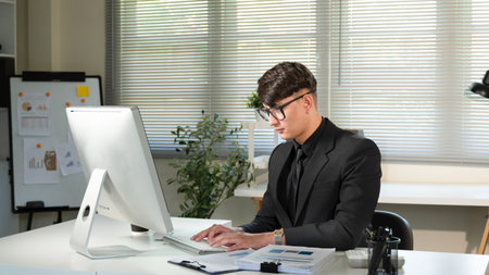 A man in a suit is sitting at a desk in front of a computer monitor. He is wearing glasses and he is...