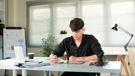A man is sitting at a desk with a laptop and a stack of papers. He is writing on the papers and he...