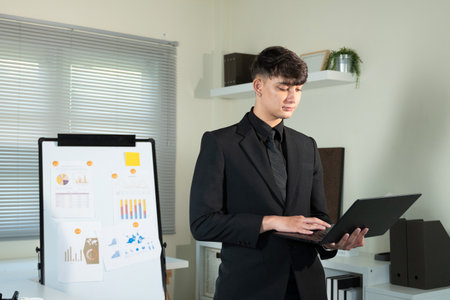 A man in a suit is holding a laptop computer. He is wearing a black tie and he is typing on the laptop..の写真素材