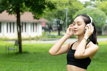 Portrait of happy beautiful Asian teenage girl wearing the headphones in outside green garden. Joyful woman enjoys relaxing and listening to music at outdoor public green park surrounded by nature..の写真素材