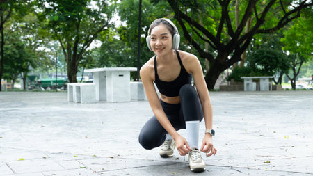Young asian woman in sportswear and headphone tying a shoelace before exercise or workout in park..の写真素材