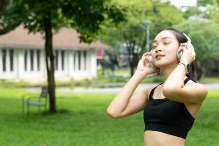 Portrait of happy beautiful Asian teenage girl wearing the headphones in outside green garden. Joyful woman enjoys relaxing and listening to music at outdoor public green park surrounded by nature..の写真素材