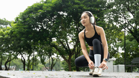 Young asian woman in sportswear and headphone tying a shoelace before exercise or workout in park..の写真素材