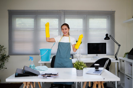 A woman in a yellow apron is cleaning a desk with a spray bottle and a bucket. She is happy and...