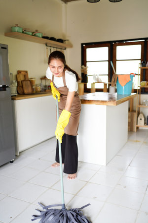 A woman is cleaning a kitchen floor with a mop. She is wearing a apron and gloves