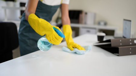 Young asian woman wearing yellow gloves and an apron, smiling while cleaning a white table with...