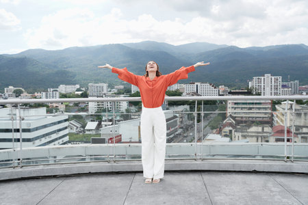 A woman in an orange shirt and white pants stands on a balcony overlooking a city. She is smiling and she is enjoying the view.の写真素材