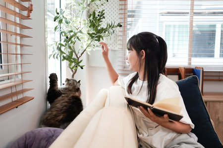 Beautiful woman with cute cat reading book near window at home.