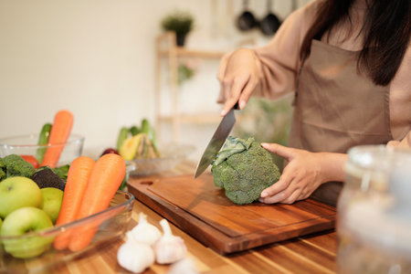 A woman is cutting a broccoli on a wooden cutting board. The kitchen is well-stocked with various fruits and vegetables, including apples, carrots.の写真素材