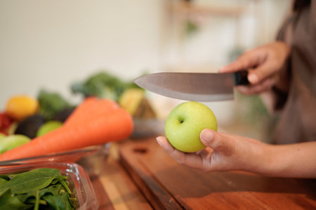 A woman wearing an apron is cutting a green apple with a knife surrounded by fresh ingredients on a...
