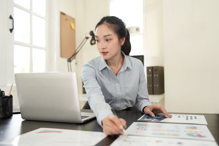 A businesswoman in a modern office setting is focused while working on a laptop and reviewing...