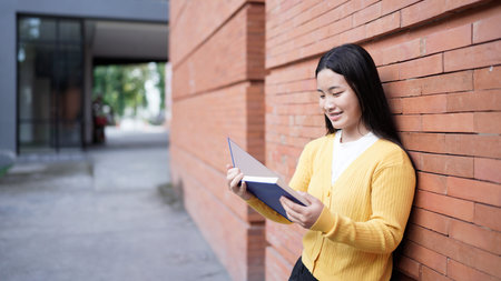 Portrait of Asian teen girl reading book on brick wall background. Beautiful young teenager student...