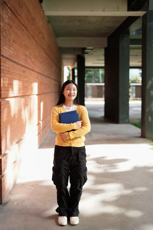 A young woman wearing a yellow sweater and black pants is holding a blue book. She is smiling and...