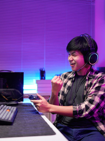 Happy asia guy gamer wear headphone participation play video game colorful neon lights computer in living room at night modern house. Esport streaming game online, Home quarantine activity concept..の写真素材