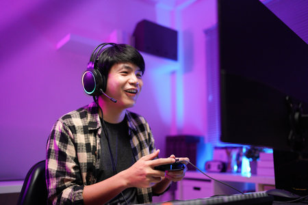 Attractive Asian male gamer enjoys playing online games on his computer feeling happy while live...