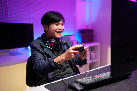 Happy asia guy gamer wear headphone participation play video game colorful neon lights computer in living room at night modern house. Esport streaming game online, Home quarantine activity concept..の写真素材