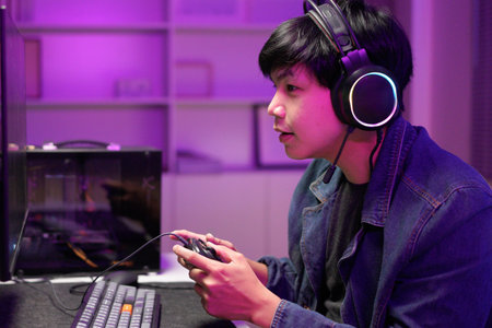 Attractive Asian male gamer enjoys playing online games on his computer feeling happy while live streaming a cyber tournament from home.の写真素材