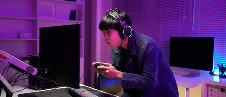 Attractive Asian male gamer enjoys playing online games on his computer feeling happy while live streaming a cyber tournament from home.の写真素材