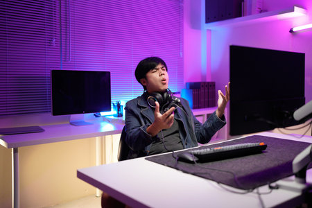 Emotional angry mad furious talking in microphone video game loss addiction asian guy male man streamer gamer wearing headphones playing virtual battle lost fail internet computer at night home.の写真素材