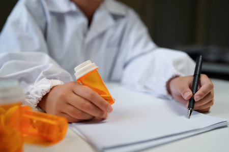 Woman doctor reviews medication bottle while writing notes in a sunlit clinic room suggesting a professional medical workplace setting..の写真素材
