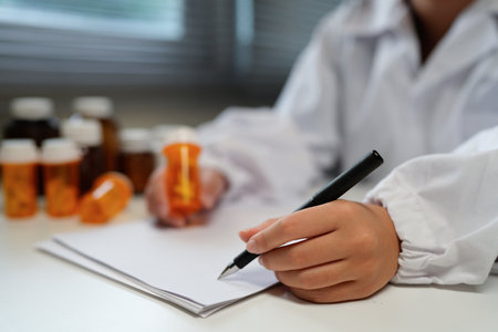 Woman doctor reviews medication bottle while writing notes in a sunlit clinic room suggesting a professional medical workplace setting..の写真素材