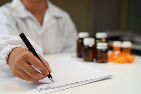 Close-up of a doctor or pharmacist's hands writing a prescription on a clipboard with medicine bottles in the background.の写真素材