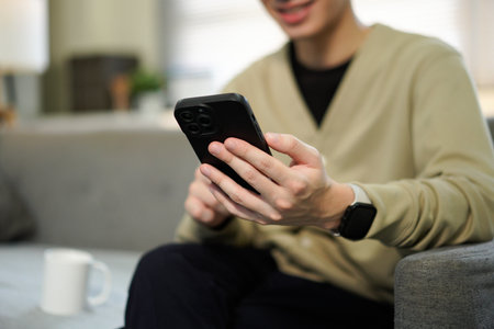 Close up of a man holding a smartphone and typing on the screen while sitting on a couch in a living room, for business, communication and technology concept..の写真素材