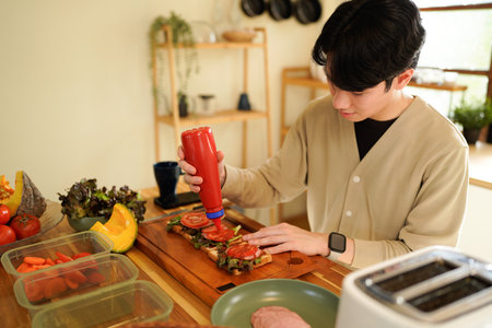 A man is making a sandwich with a red condiment. The sandwich is on a wooden cutting board in kitchenの写真素材