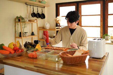 A man is making a sandwich with a red condiment. The sandwich is on a wooden cutting board in kitchen.の写真素材