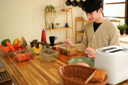 A man is preparing a sandwich on a wooden cutting board. The kitchen is well-stocked with various food items.の写真素材