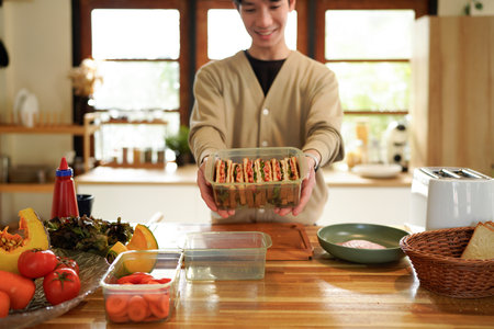 A man is holding a container of sandwiches in kitchen. The sandwiches are in a plastic container and are cut into halves..の写真素材
