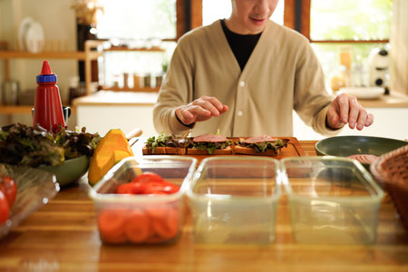 Man Making Sandwiches at bright and minimalist Counter with Fresh Greens, ham. Asian man cooking healthy foods in cozy kitchen in morning at home..の写真素材
