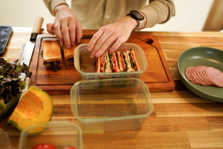 A man is preparing sandwiches in a kitchen. He is using a knife to cut the bread and is placing the...