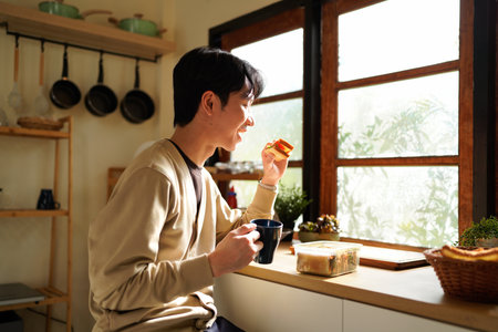 A man is eating a sandwich and drinking coffee near the window in the morning. Scene is relaxed and casual.の写真素材