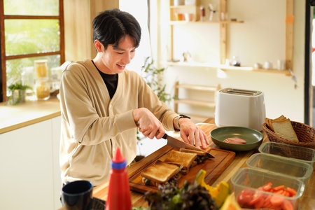 A man is preparing a sandwich on a wooden cutting board. The kitchen is well-stocked with various food items.の写真素材