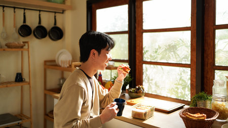 A man is eating a sandwich and drinking coffee near the window in the morning. Scene is relaxed and casual.の写真素材
