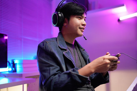 Attractive Asian male gamer enjoys playing online games on his computer feeling happy while live streaming a cyber tournament from home.の写真素材