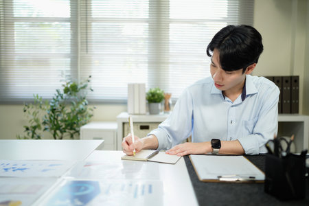 Businessman analyzing data (graphs/charts) on a document, taking notes with a pen. Office setting, professional work.の写真素材